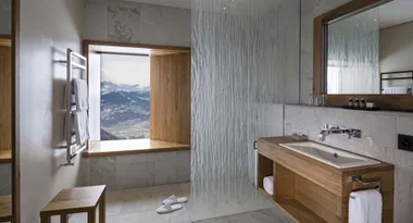 Modern bathroom with mountain view, wood accents, glass partition, towel rack, and slippers on tiled floor.