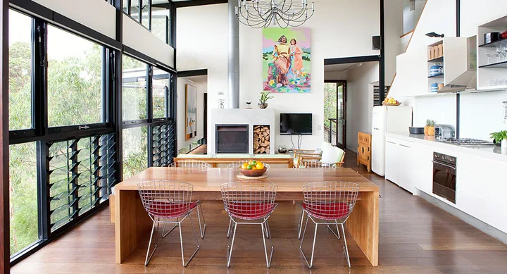 Modern open-plan living area with large windows, wooden dining table, wire chairs, and a colorful artwork above the fireplace.