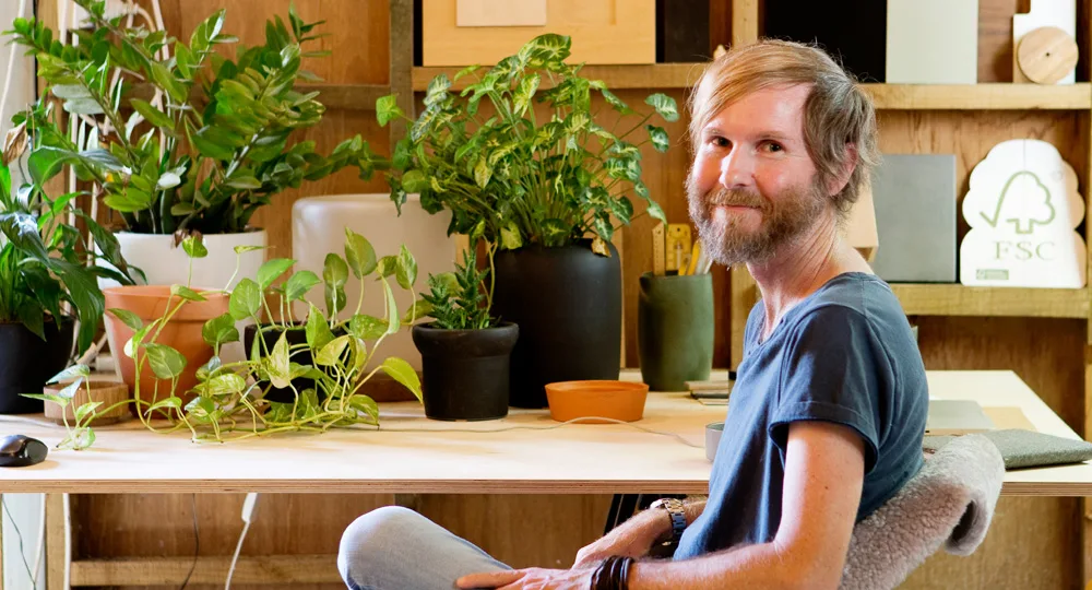 A person with a beard sits at a desk surrounded by potted plants, smiling and wearing a blue shirt.