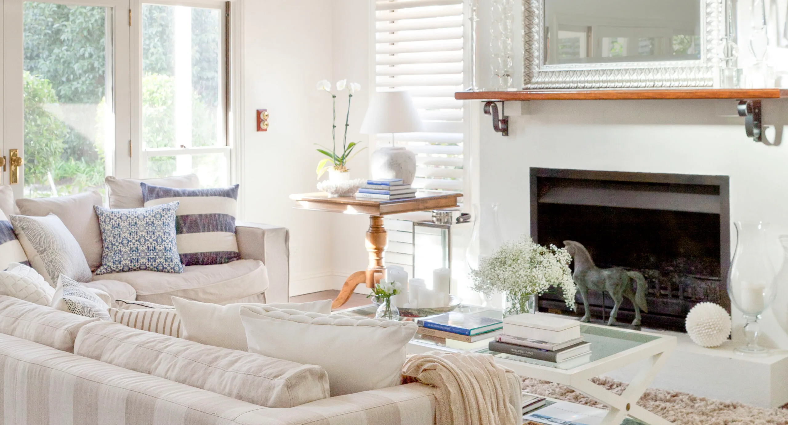 Bright living room with white sofas, blue cushions, a mirror, fireplace, and a wooden table holding books and an orchid.