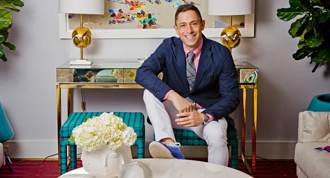 Man sitting on a teal bench in a stylish room with gold accents, floral decor, and artwork on the wall.