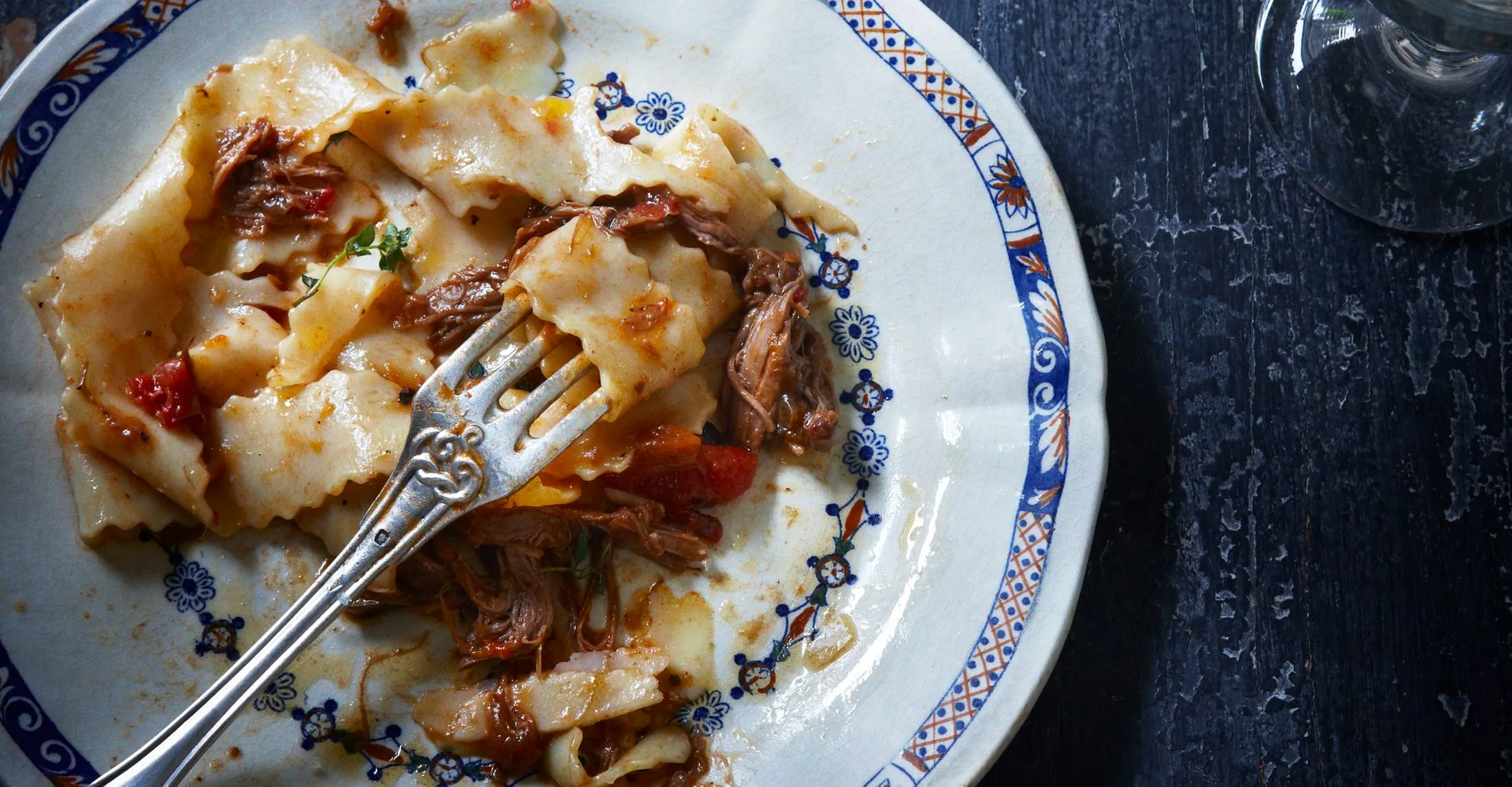 Plate of pasta with slow-cooked meat, tomato sauce, and a fork, on a white and blue decorative plate.