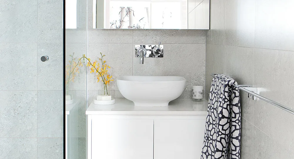 Modern bathroom with gray tiles, white countertop, vessel sink, wall-mounted faucet, mirror, plant, and patterned towel.