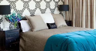 Elegant bedroom with patterned wallpaper, brown and white pillows, a turquoise blanket, and bedside lamps.