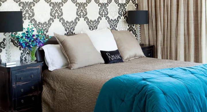 Elegant bedroom with patterned wallpaper, brown and white pillows, a turquoise blanket, and bedside lamps.