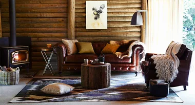 Cozy log cabin living room with leather sofa, armchair, wood stove, rustic decor, and sunlight pouring in.