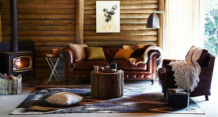 Cozy log cabin living room with leather sofa, armchair, wood stove, rustic decor, and sunlight pouring in.