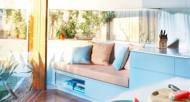 Sunny kitchen nook with orange and blue cushions, books, and a window view of a garden with greenery.