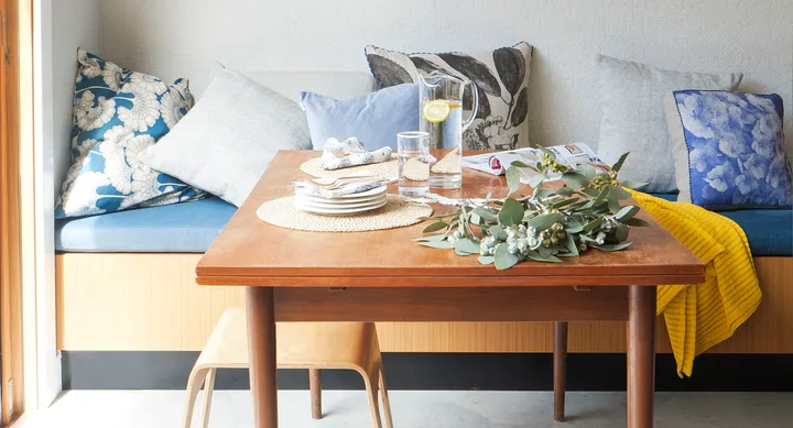 Cozy dining nook with a wooden table, floral pillows, a yellow blanket, and glass pitcher with lemon slices.