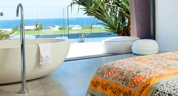 Luxurious bedroom with a large bathtub, colorful bedspread, and ocean view through glass doors.