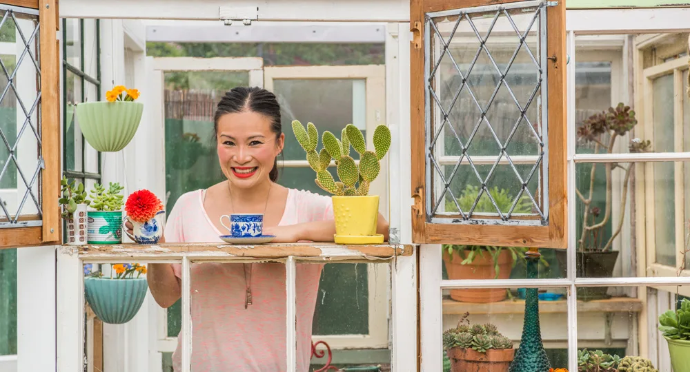 Woman smiling at a window in a greenhouse with colorful plants and succulents.