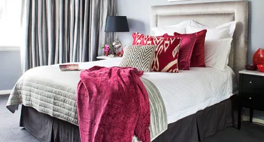 Elegant bedroom with gray and red accents, featuring a plush bed with patterned pillows and a maroon throw blanket.