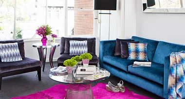 Living room with blue sofa, patterned cushions, pink rug, glass table, and floral decorations by the window.