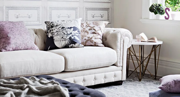 Cream sofa with patterned cushions, beside a round table holding a book and glass, in a bright room with decorative paneling.