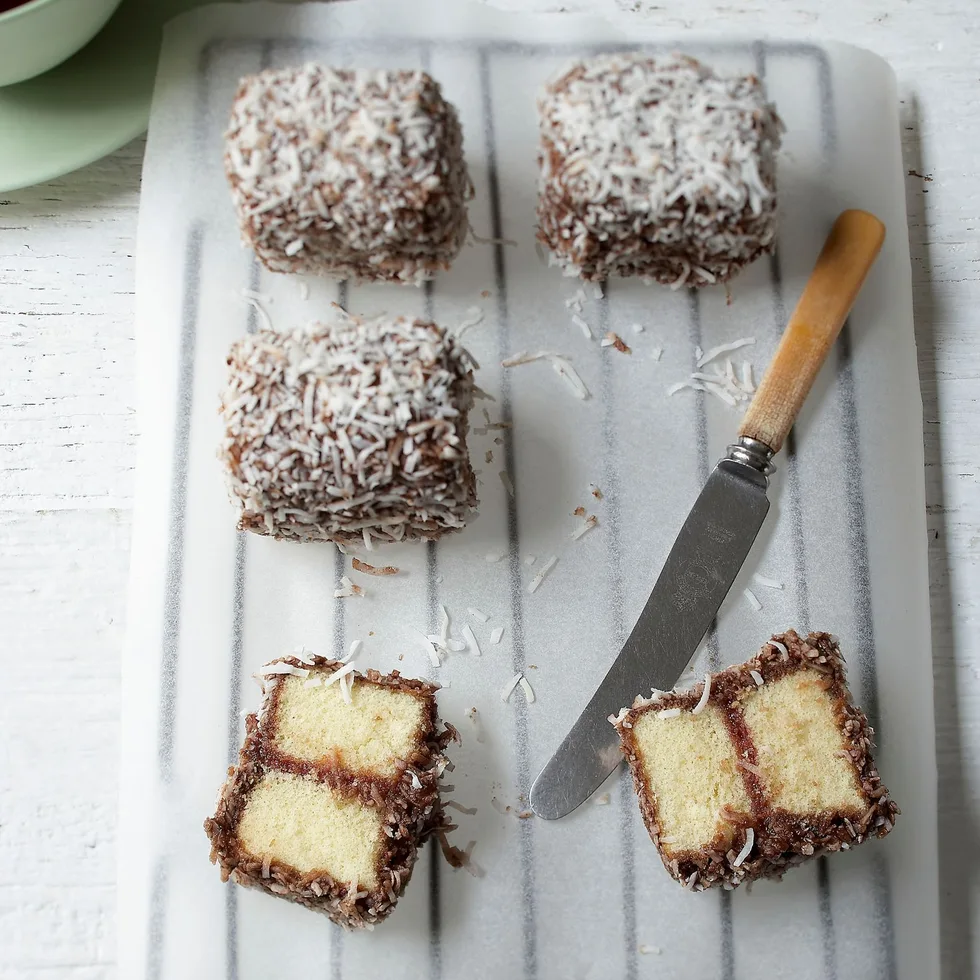 Lamingtons coated with chocolate and coconut on parchment paper, accompanied by a knife and a cup of tea.