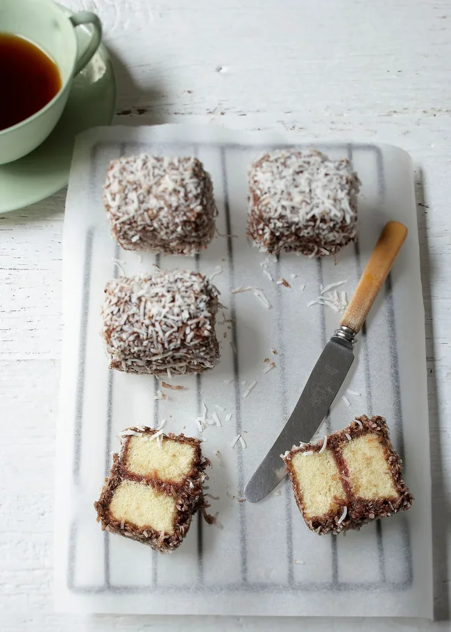 Lamingtons coated with chocolate and coconut on parchment paper, accompanied by a knife and a cup of tea.