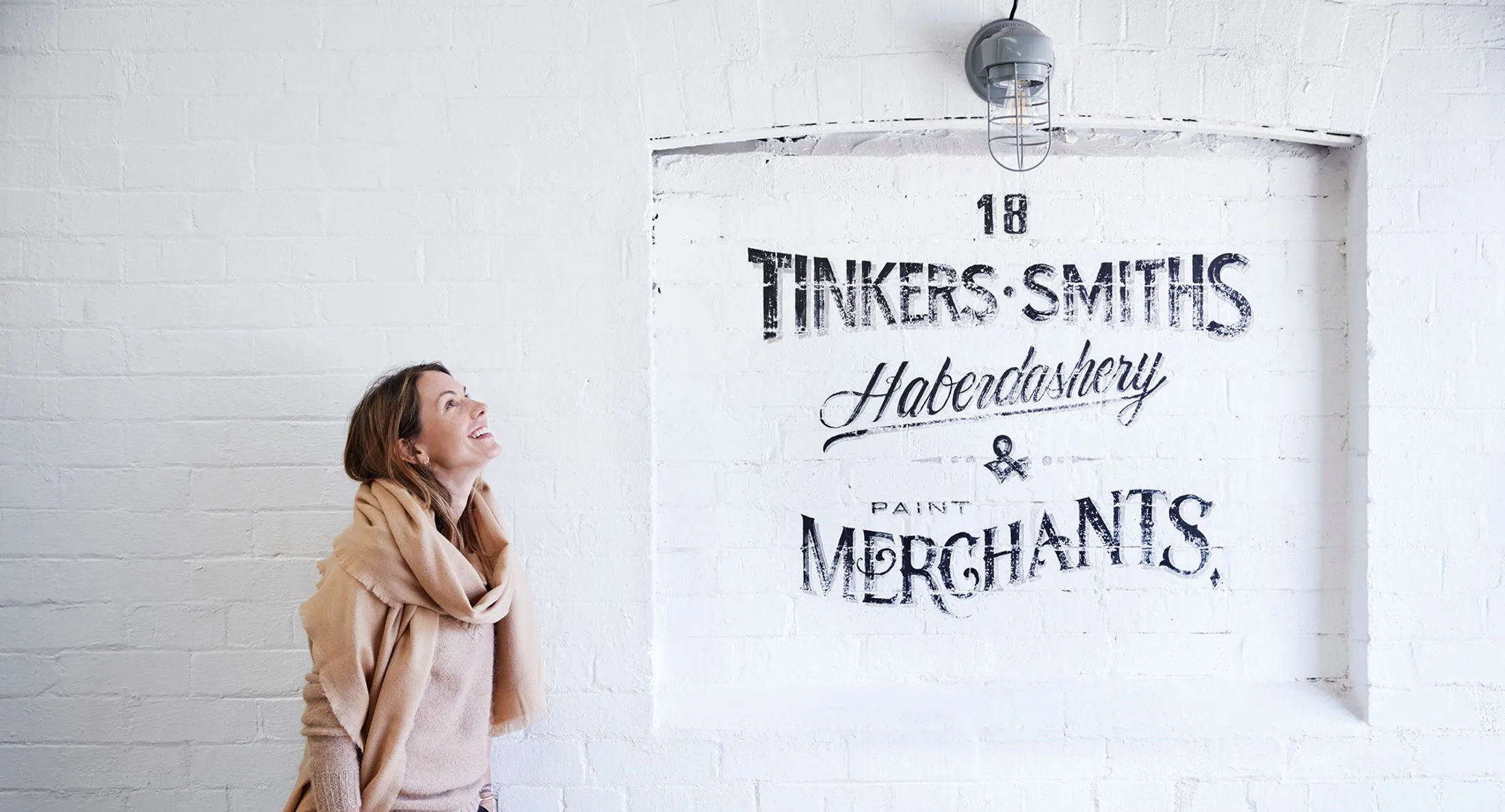 Woman in a beige scarf looking at wall art reading "Tinkers Smiths Haberdashery & Paint Merchants."