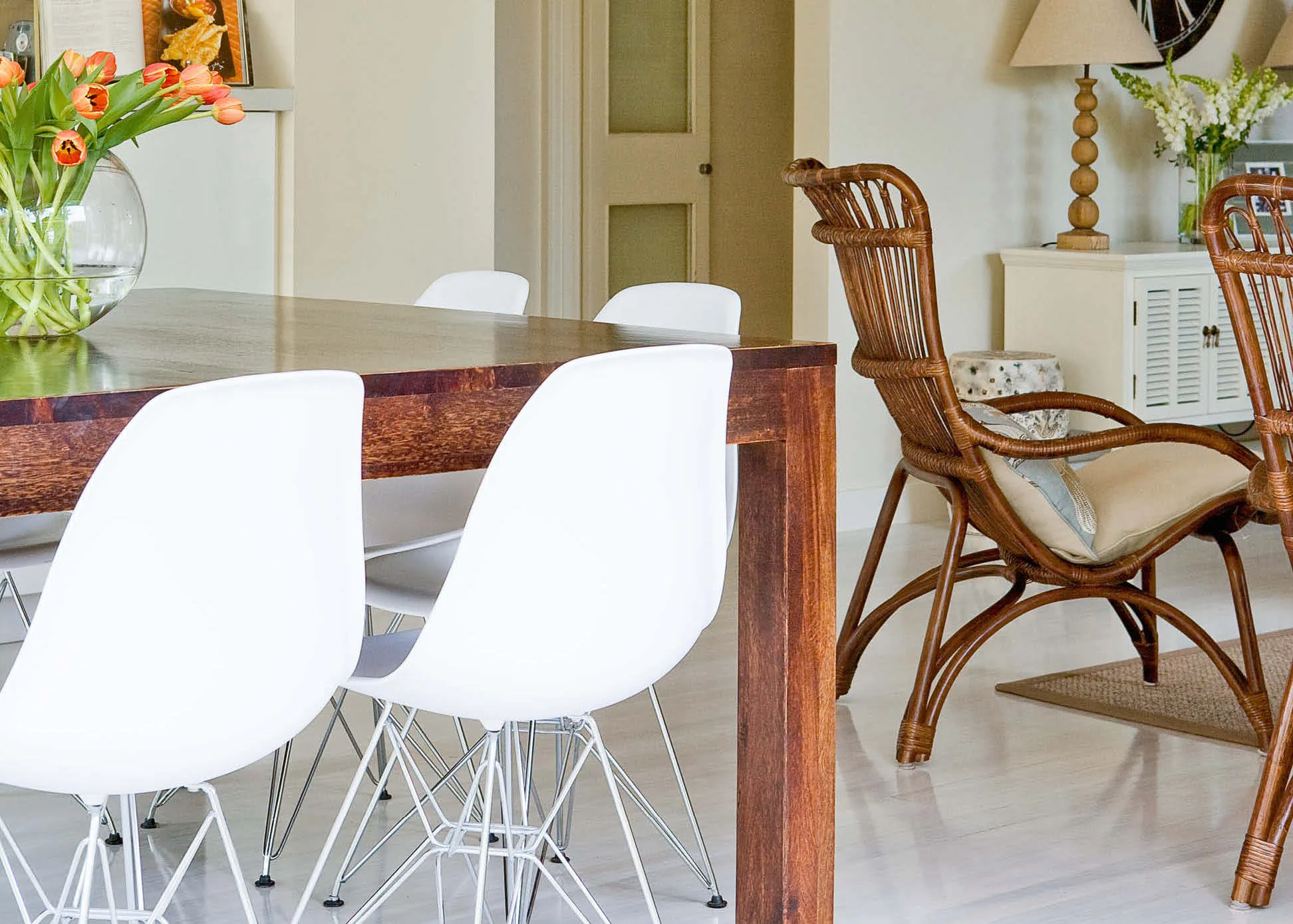 Dining room with a wooden table, white chairs, a wicker chair, a vase of orange tulips, and a lamp on a sideboard.