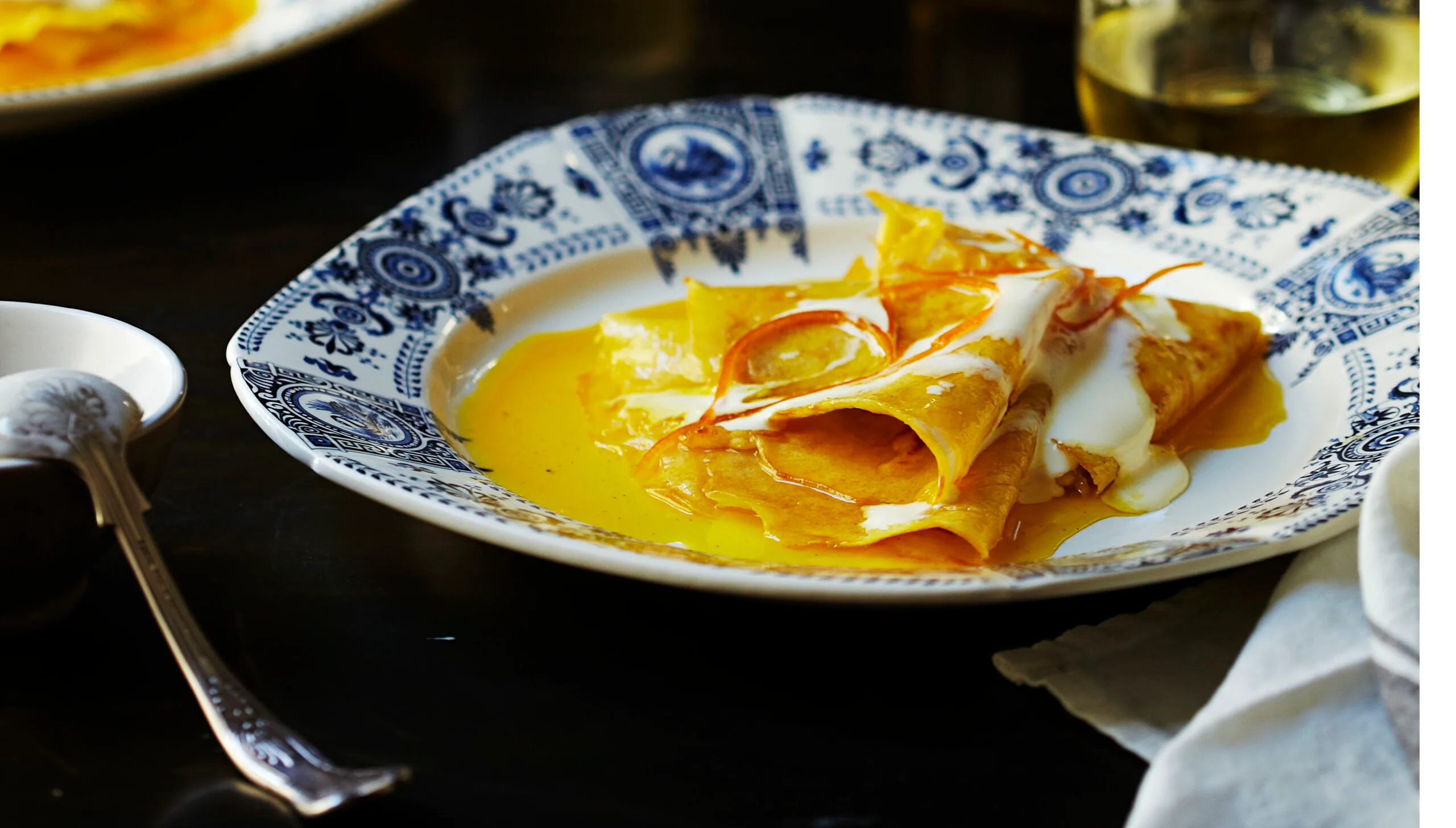 Crepes Suzette with orange sauce and cream on a patterned plate, set on a dark table with a spoon nearby.