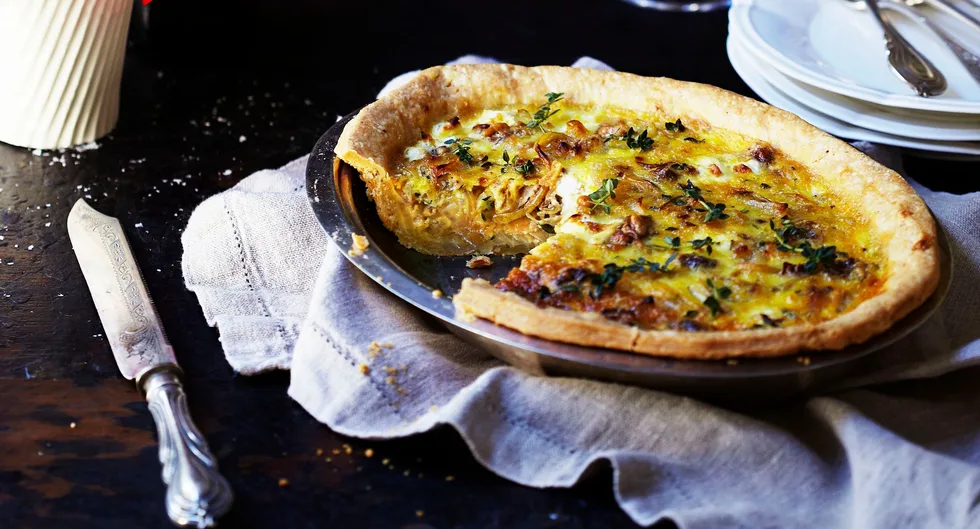 A savory quiche with a golden crust, garnished with herbs, on a cloth napkin beside an ornate silver knife.
