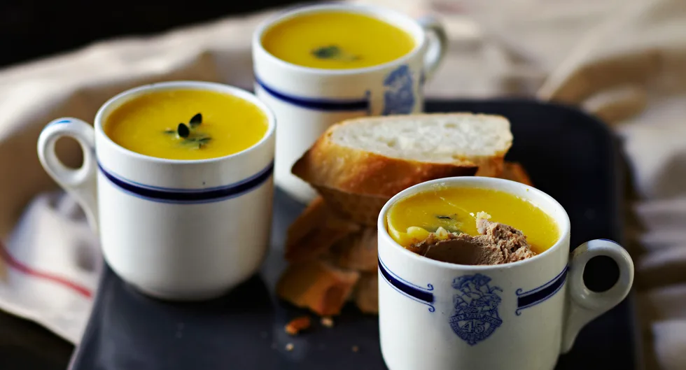 Mugs of pumpkin soup garnished with seeds, served with slices of rustic bread.