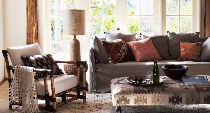 Cozy living room with a sofa, colorful cushions, rustic chair, large window, and decorative items on a patterned floor.