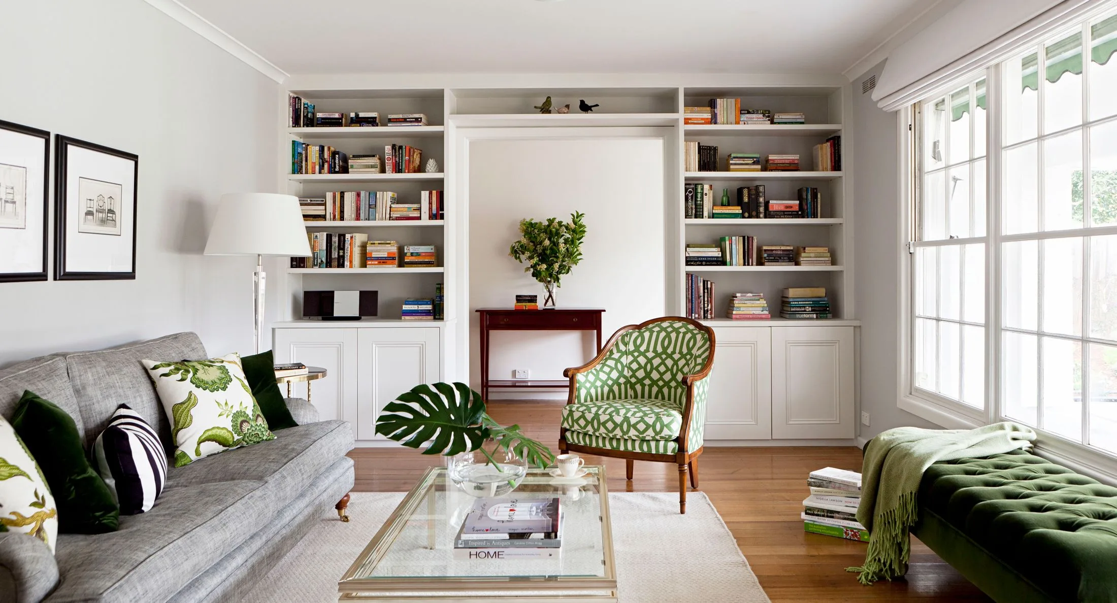 Bright living room with a gray sofa, patterned green chair, bookshelves, and a large window.