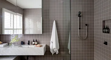 Modern bathroom with grey tiles, a marble sink, toiletries, a towel, and a glass-enclosed shower area.