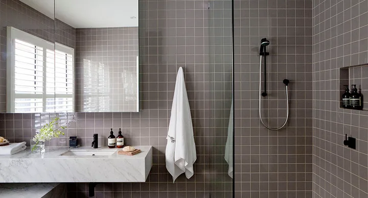 Modern bathroom with grey tiles, a marble sink, toiletries, a towel, and a glass-enclosed shower area.