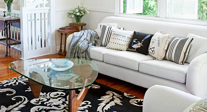 Bright living room with a white sofa, patterned cushions, a glass coffee table, and a floral black and white rug.
