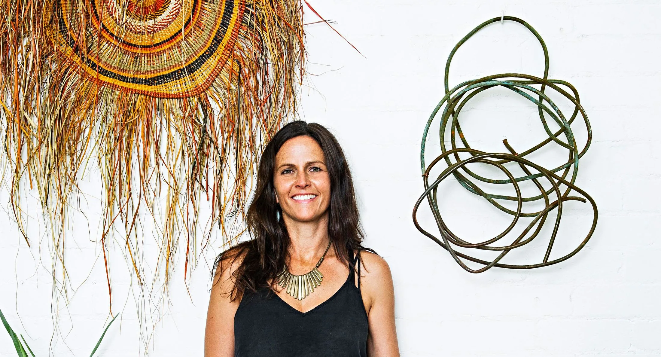 Woman with long hair smiling, standing in front of woven art and metal sculpture on a white wall.