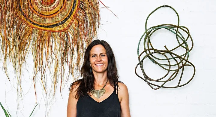 Woman with long hair smiling, standing in front of woven art and metal sculpture on a white wall.