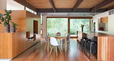 Modern kitchen and dining area with wooden floors, large table, white chairs, island, and greenery outside the window.