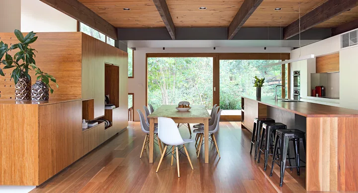 Modern kitchen and dining area with wooden floors, large table, white chairs, island, and greenery outside the window.