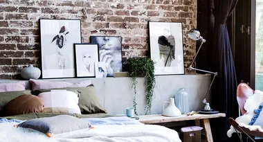 Cozy bedroom with a brick wall, framed art, a desk lamp, pillows, and plants.