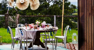 Outdoor table setting with white chairs, tablecloth, flowers, and decorative paper fans, in a green garden setting.