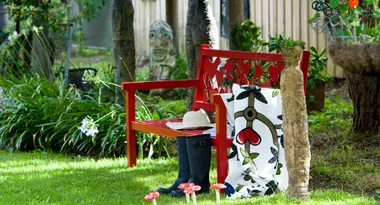 Red bench in a garden with boots, a hat, a patterned blanket, and decorative mushrooms on the grass.