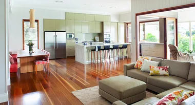 Modern open-plan kitchen and living area with wooden floors, olive cabinets, bar stools, and floral cushions on sofas.