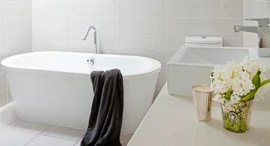 Modern bathroom with white bathtub, dark towel draped over its side, sink, flowers, and a candle on counter.