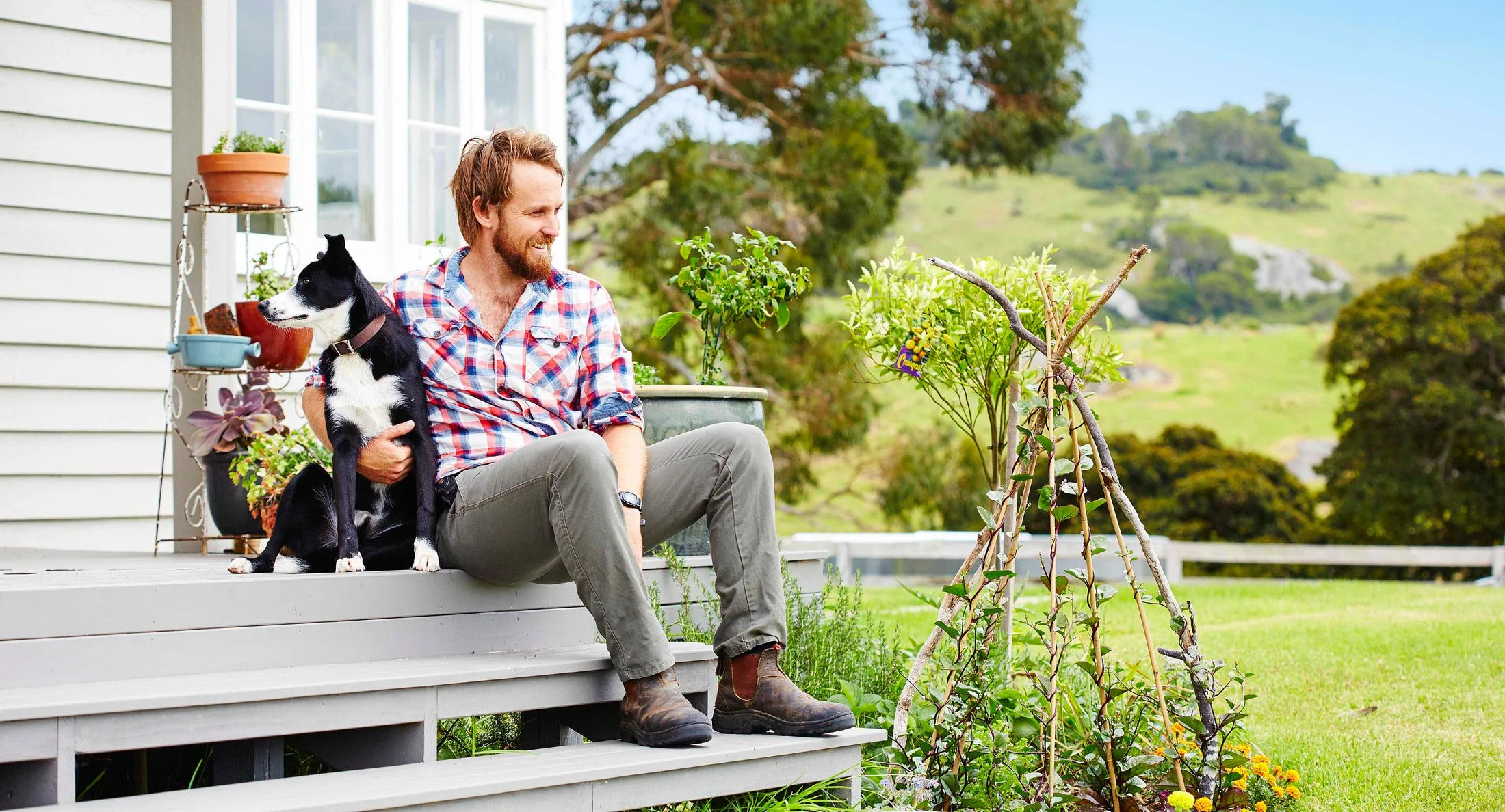 Man in plaid shirt sitting on steps with a black and white dog, garden plants, and green landscape in the background.
