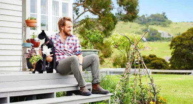 Man in plaid shirt sitting on steps with a black and white dog, garden plants, and green landscape in the background.