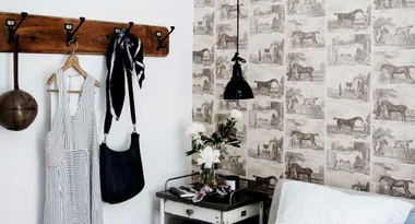 Cozy room with horse-patterned wallpaper, black lamp, a coat hanger with clothes, and a small table with flowers.
