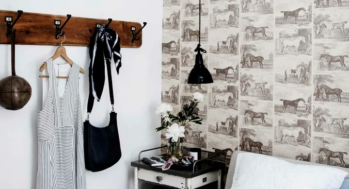 Cozy room with horse-patterned wallpaper, black lamp, a coat hanger with clothes, and a small table with flowers.