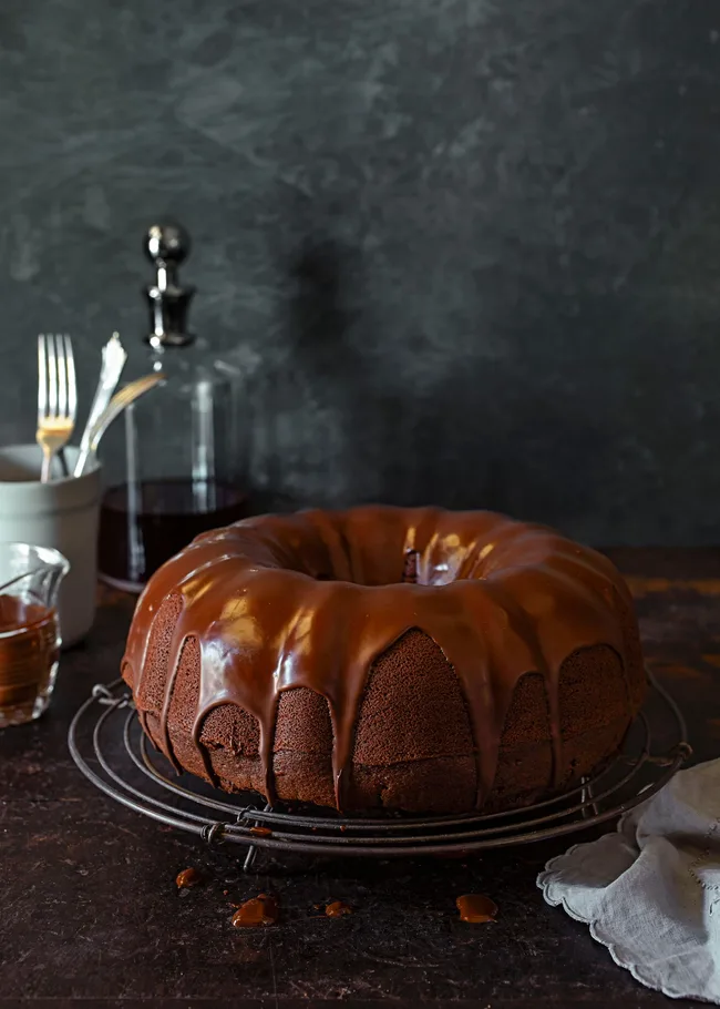 Chocolate bundt cake with glossy glaze on a cooling rack, set in a moody kitchen scene.