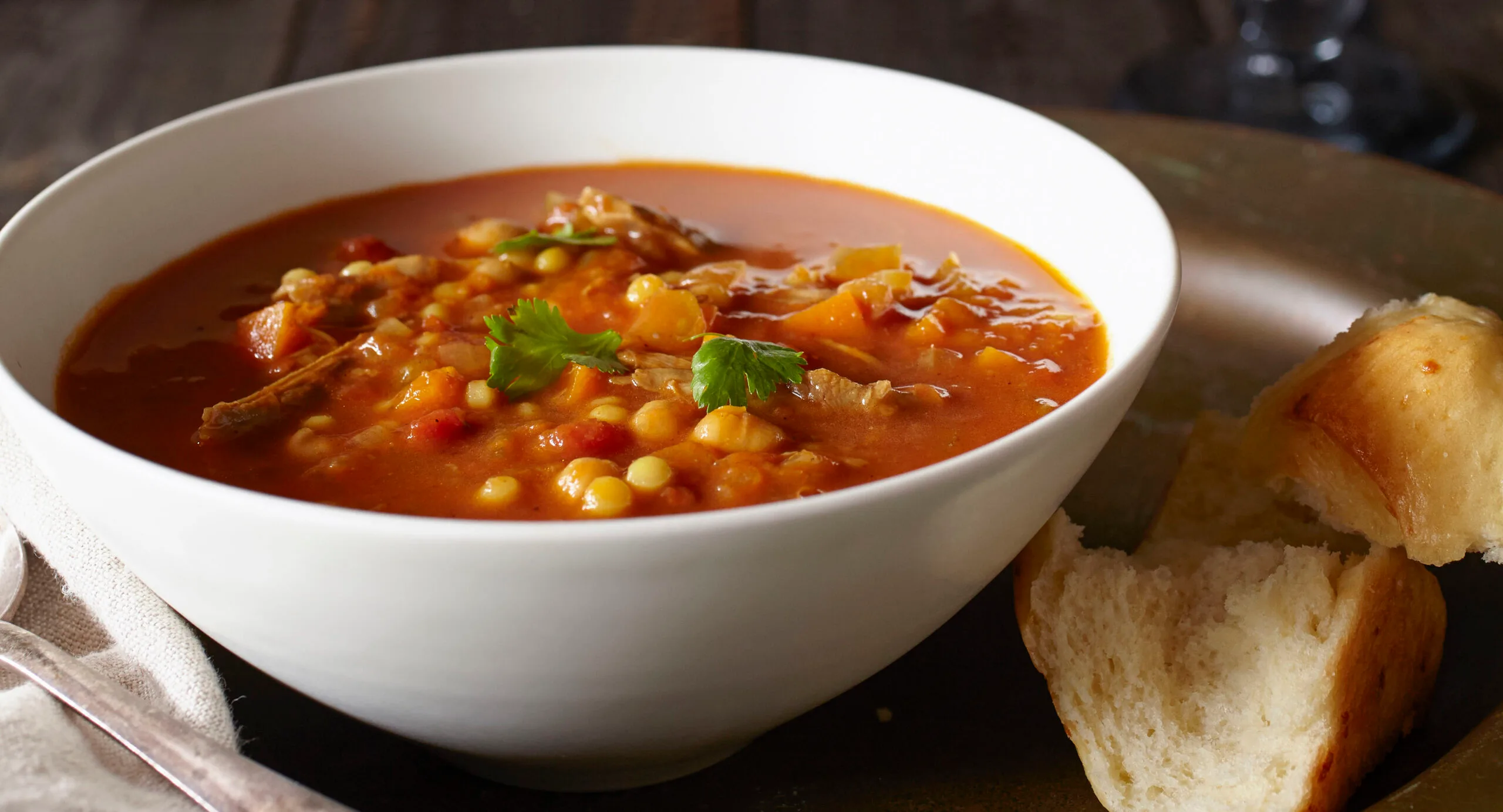 Bowl of hearty soup with vegetables and herbs, beside a piece of bread on a plate.