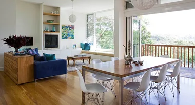 Modern open-plan living room and dining area with wooden flooring, blue sofa, contemporary art, and large windows.