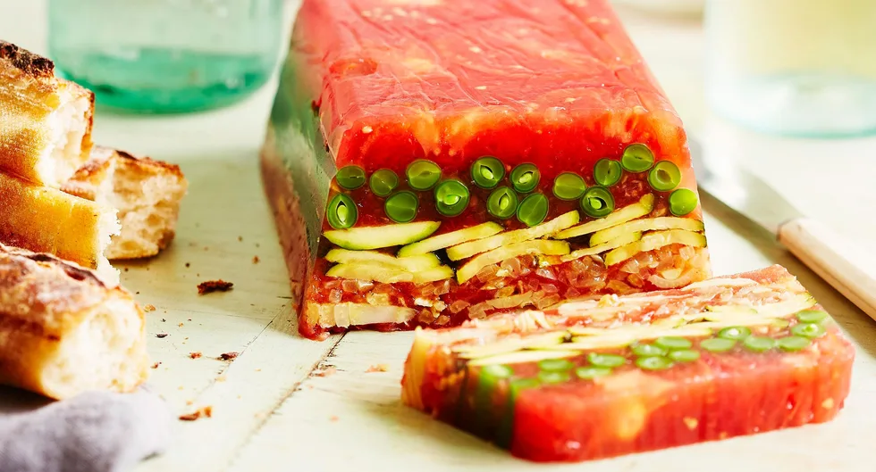 Layered vegetable terrine with tomatoes and peas, served on a light wooden table with rustic bread and knife.