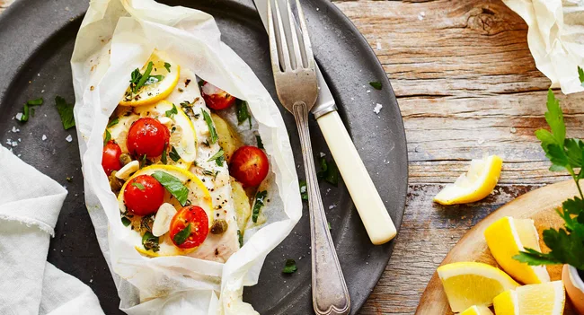 Fish baked in parchment with lemon, cherry tomatoes, and herbs, served on a plate with a fork and knife.