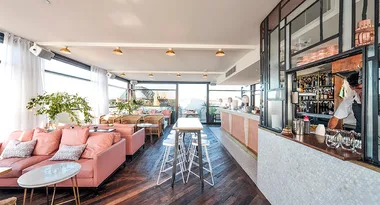 Bright rooftop bar with pink seating, marble tables, and a bartender behind the counter, overlooking a cityscape.