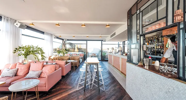 Bright rooftop bar with pink seating, marble tables, and a bartender behind the counter, overlooking a cityscape.